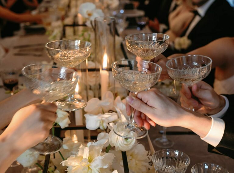 A group of people raise champagne glasses in a toast at a long, elegantly decorated banquet table adorned with white flowers and candles. The scene is festive, and everyone appears to be dressed in formal attire.