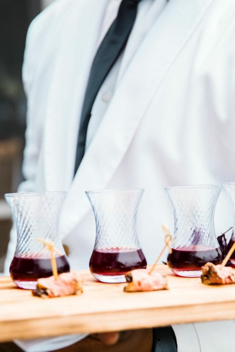 A person in a white jacket with black tie is holding a wooden tray with five glasses of red wine and small skewered appetizers. The background is blurry, focusing attention on the tray and its contents.