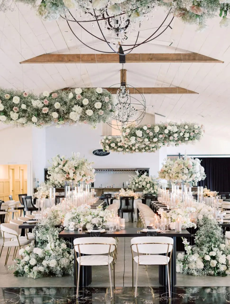 A luxurious wedding reception hall is elegantly decorated with abundant floral arrangements, featuring white and blush flowers. Modern white chairs surround tables adorned with candles and flowers. Flower installations hang from the ceiling.