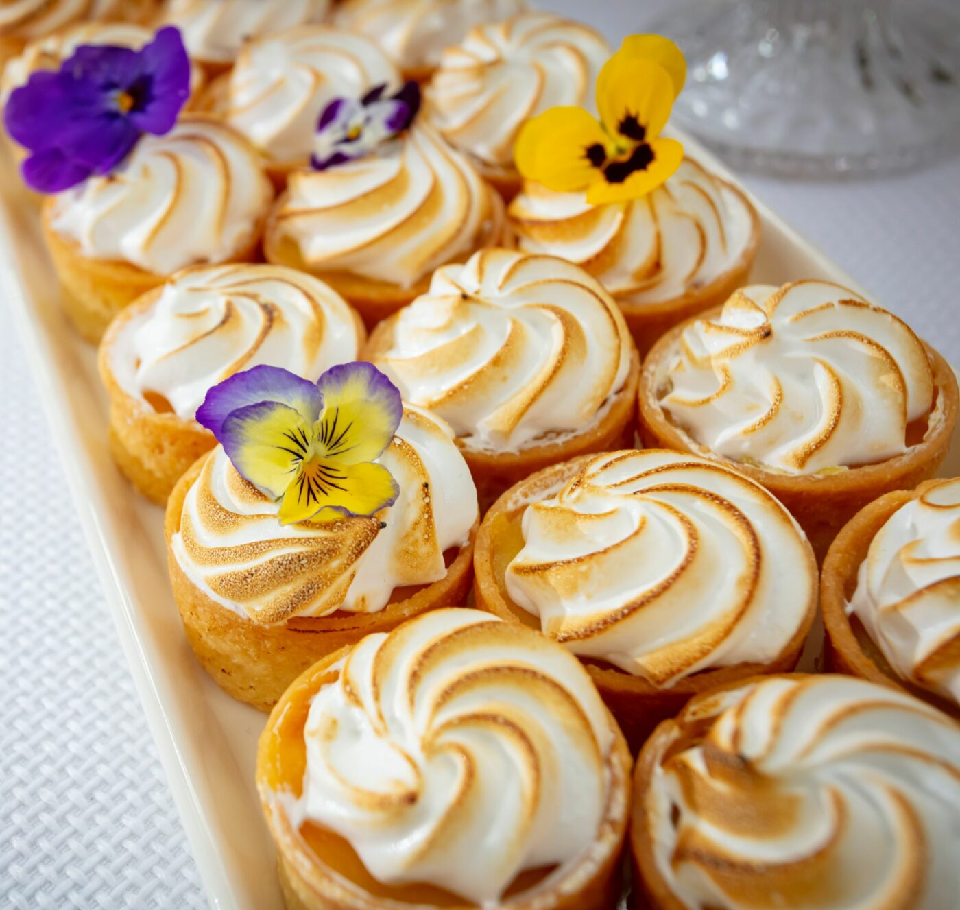 A rectangular white tray displays a dozen small tarts with swirled meringue toppings. Each tart is garnished with a colorful edible flower in shades of purple, yellow, and white. The tray is on a white textured surface.