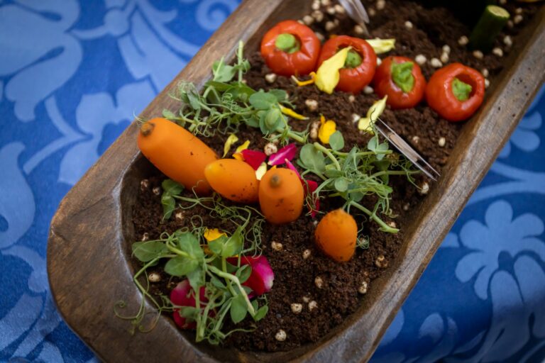 A wooden tray filled with vegetables and garnishes arranged artistically to resemble plants growing in soil.