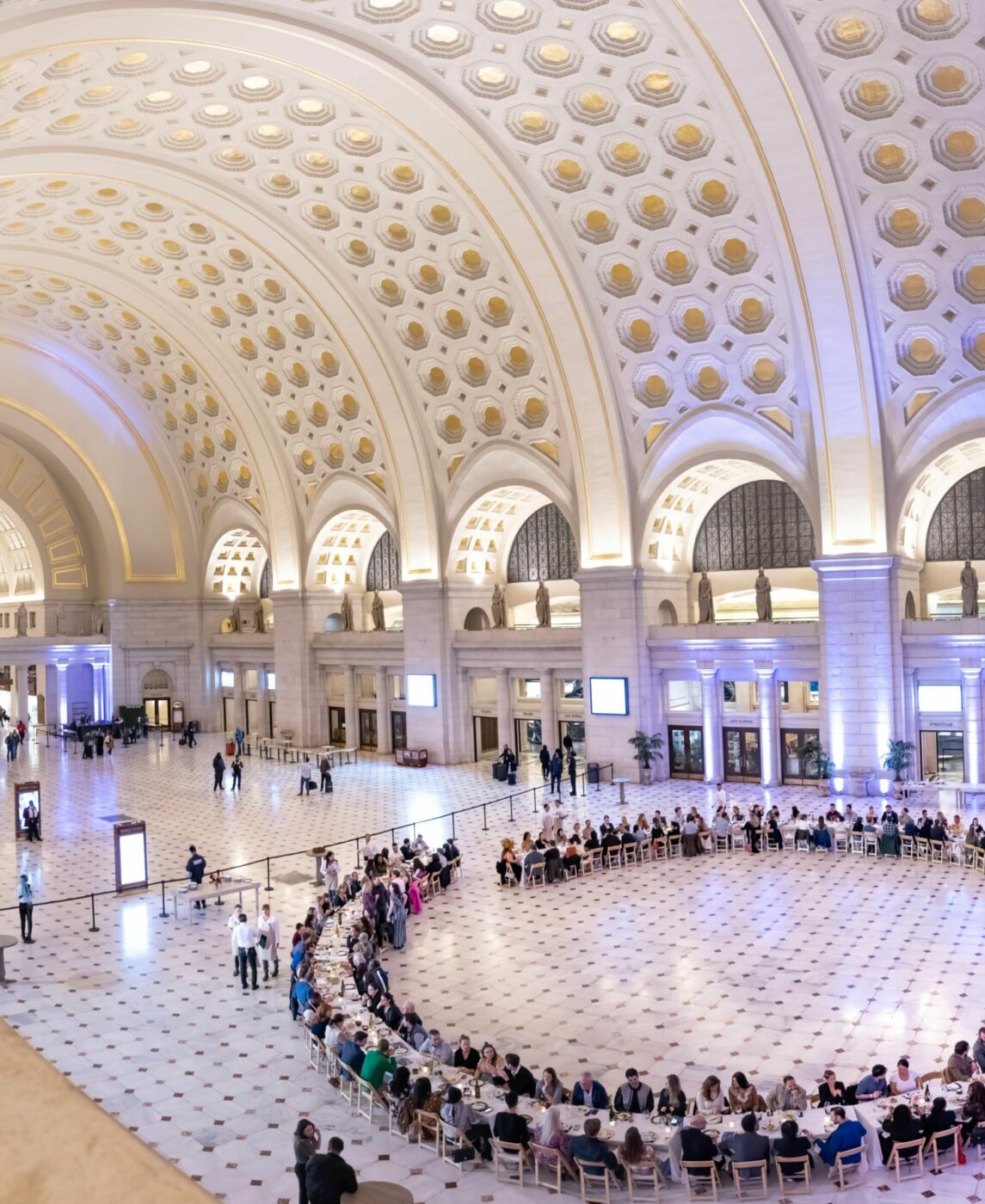 A large, elegant hall with a domed ceiling and ornate arches hosts a gathering where numerous people are seated in a circular arrangement at tables.