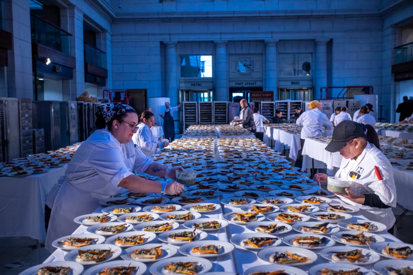 Several chefs in a spacious corridor of Union Station are meticulously plating hundreds of dishes on long tables. Each plate features a variety of gourmet food items. The scene suggests preparation for a large event or banquet.