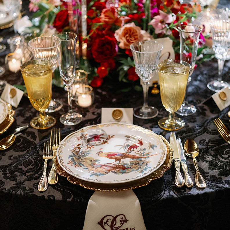 A luxurious table setting features ornate, gold-trimmed porcelain plates adorned with bird motifs. Golden cutlery and crystal glassware are neatly arranged on a black, floral-patterned tablecloth, complemented by elegant name cards and a vibrant floral centerpiece.