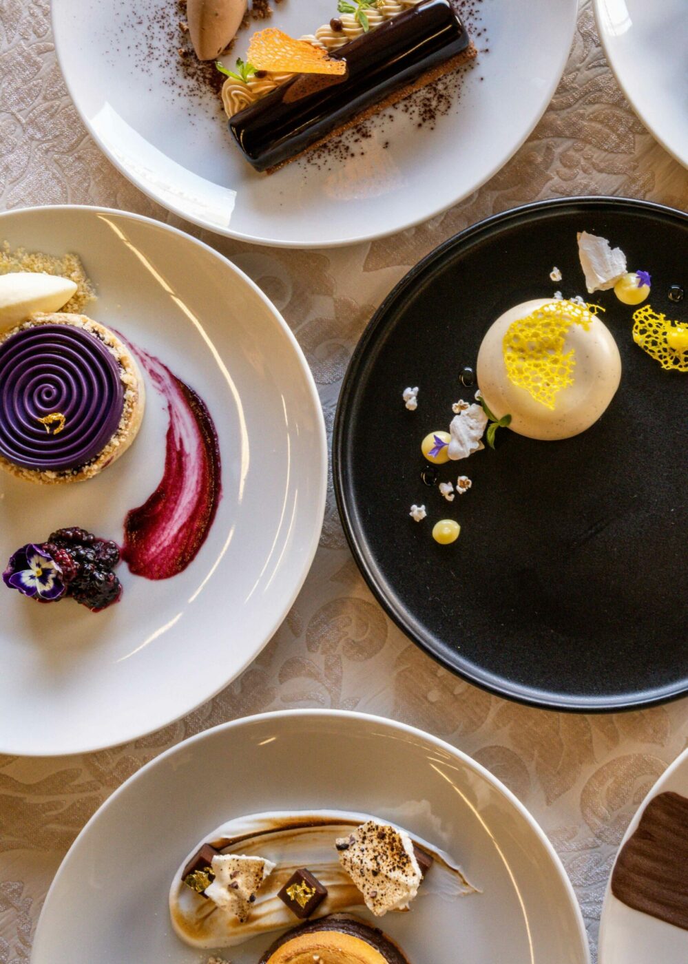 A variety of colorful gourmet desserts arranged on six white plates around one black plate, each depicting intricate presentations. The treats include cakes, mousses, and pastries adorned with fruits, edible flowers, and decorative elements, displayed on a lace tablecloth.