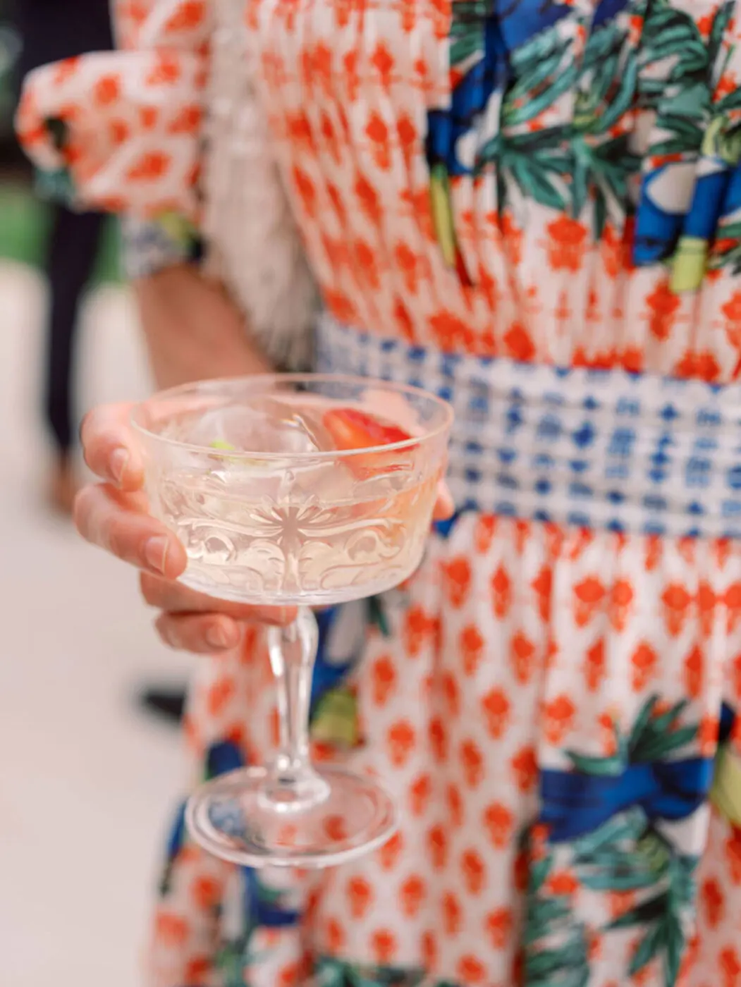 A person wearing a colorful dress holds a decorative coupe glass filled with a light-colored beverage, garnished with a slice of lime and a strawberry.