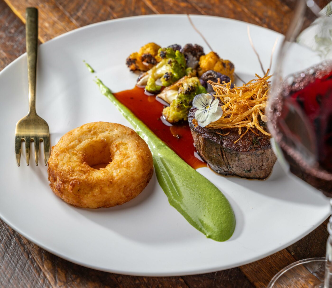 A gourmet meal on a white plate features a donut-shaped bread, a steak garnished with crispy fried onions, and vegetables, alongside a green puree. A glass of red wine is positioned to the right, partially obscuring the view of the dish. A fork is placed on the left.