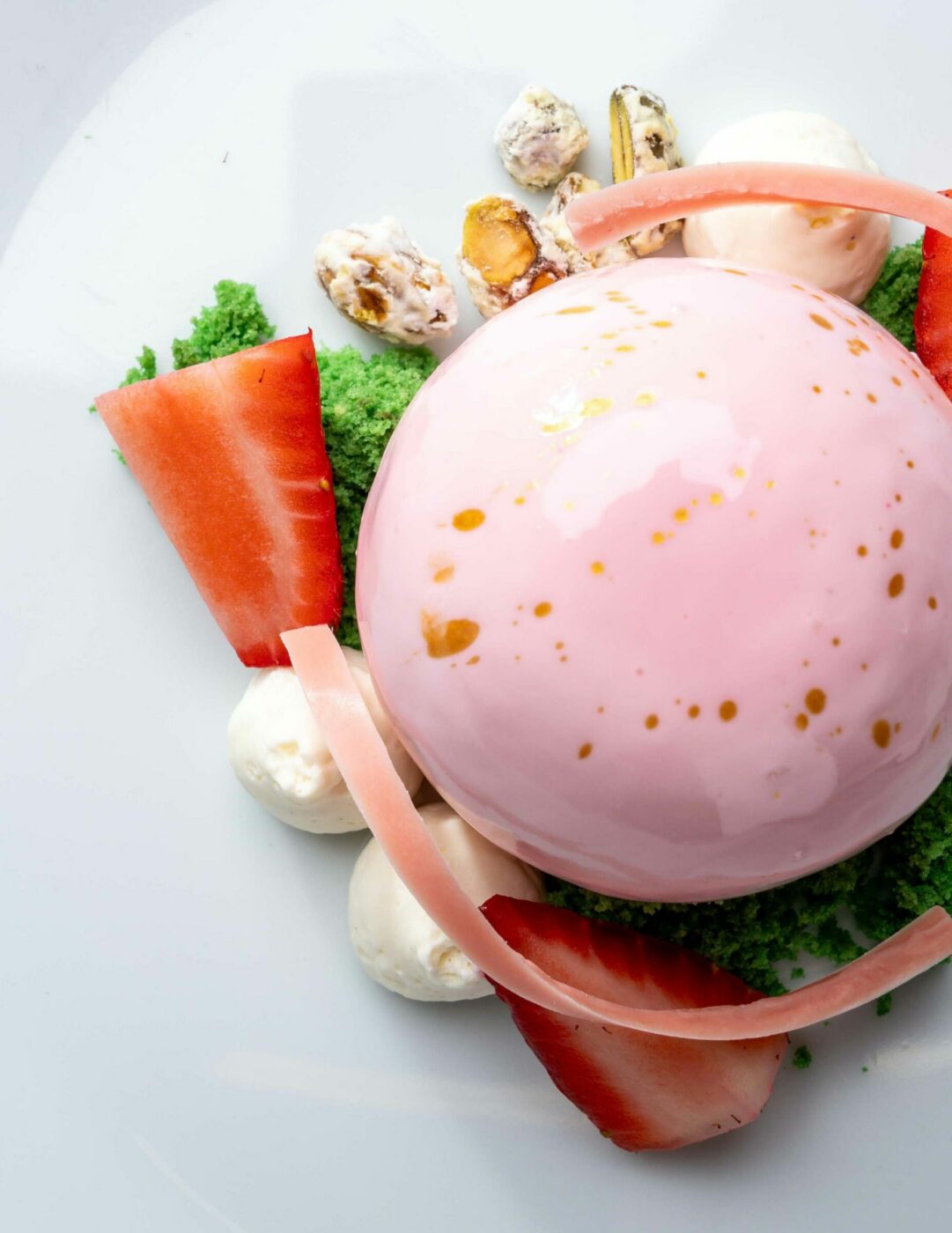 A plated dessert featuring a glossy pink dome on a white plate. It is surrounded by strawberries, cream, and garnishes, and is accompanied by a gold fork. The plate is set on a patterned golden tablecloth.