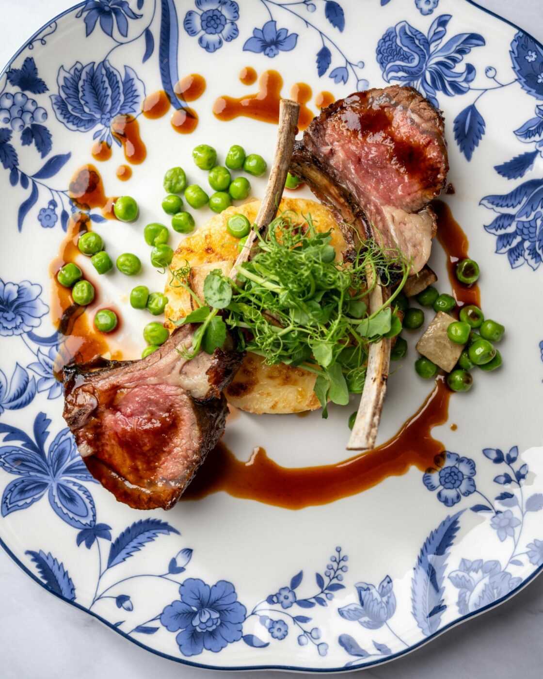 A beautifully plated dish featuring two lamb chops on a decorative blue and white plate. The lamb is garnished with a vibrant green herb, accompanied by peas and a sauce drizzle. A round, golden-brown piece of polenta lies beneath the garnished lamb.