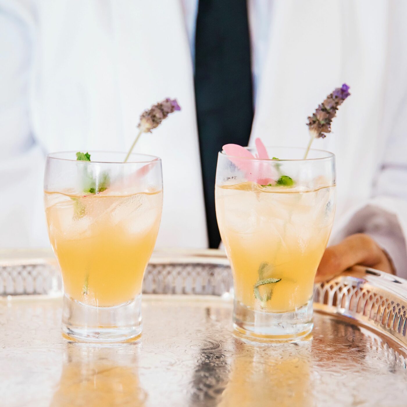 Two iced beverages garnished with lavender and flower petals on a silver tray, served by a person in a white jacket and black tie.