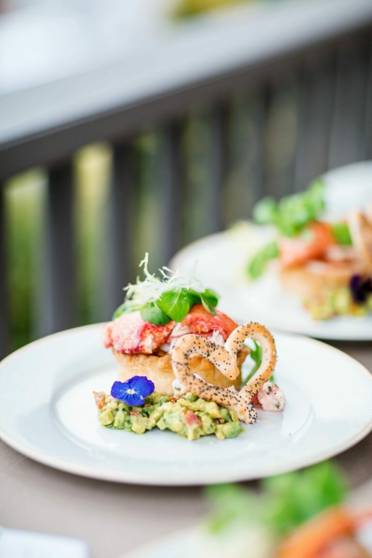 A plated gourmet dish featuring a pastry topped with a red sauce, garnished with fresh greens, and accompanied by a dollop of guacamole and a heart-shaped cracker. A purple edible flower adds a touch of color to the presentation. Other similar plates are in the background.